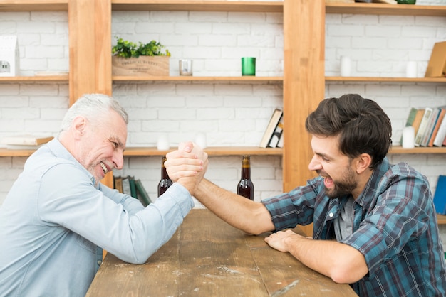 Aged man and young guy with hands clasped in arm wrestling challenge at table in room