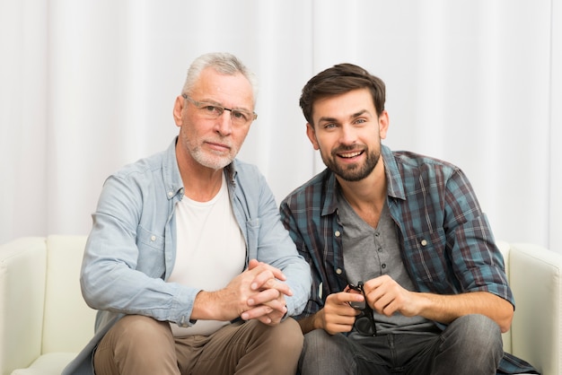 Aged man and young happy guy clasping hands on sofa