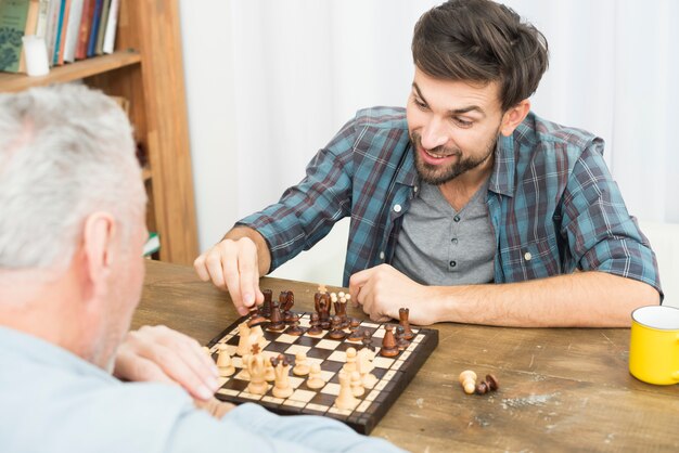 Aged man and young happy guy playing chess at table in room