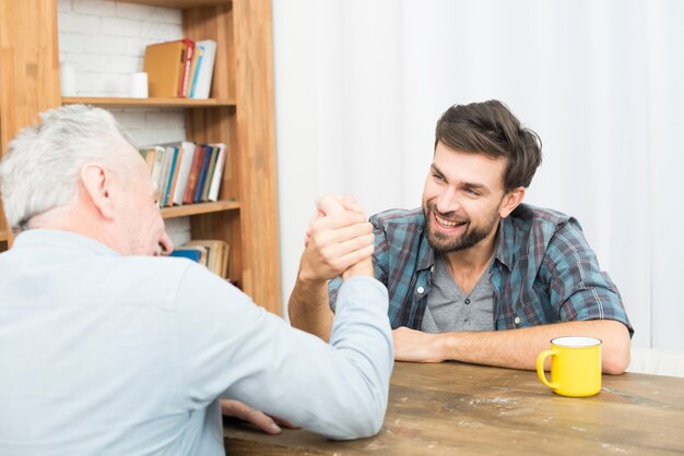 Aged man and young happy guy with hands clasped in arm wrestling challenge at table in room