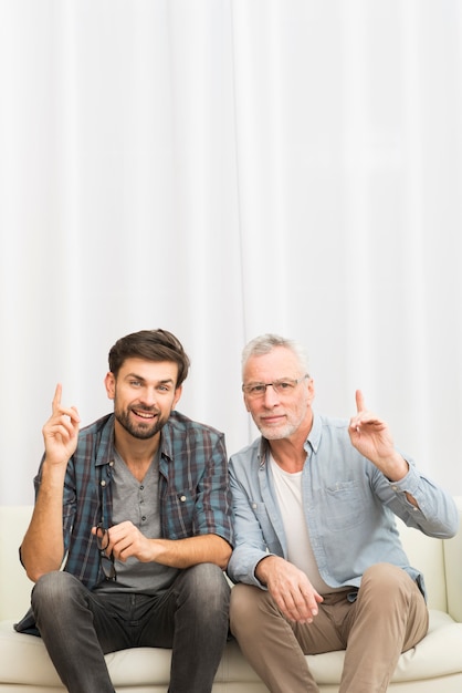 Aged man and young happy guy with upped fingers on sofa