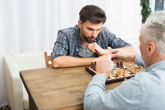 Aged man and young pensive guy playing chess at table in room