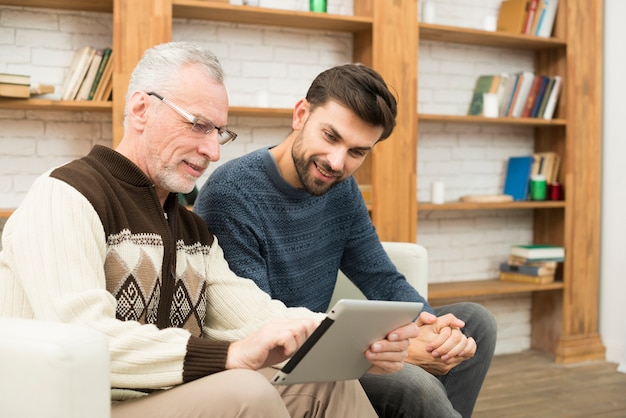 Aged man and young smiling guy using tablet on settee