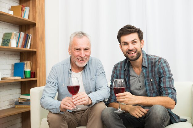 Aged man and young smiling guy with glasses of wine on settee