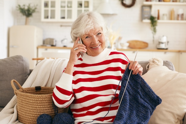 高齢者、退職、レジャー、現代技術の概念。リビングルームのソファでスカーフを編んでいる間、携帯電話で彼女の孫娘と話している白髪の美しい幸せなおばあちゃん