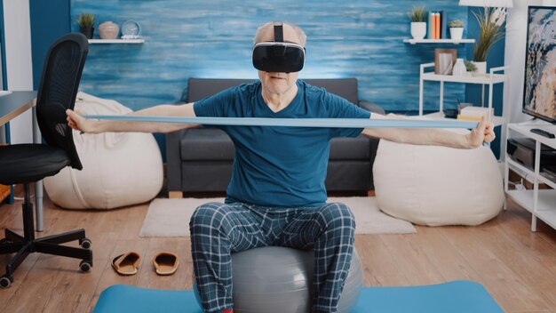 Aged person pulling resistance band and wearing vr glasses while sitting on toning ball at home. Retired man using virtual reality headset and training with stretching elastic belt.