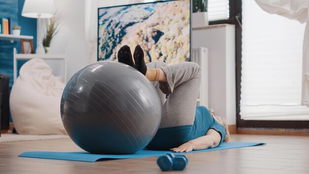 Aged person using fitness toning ball to train muscles on yoga mat. Senior woman training with gymnastics equipment to stretch legs and exercise for wellness. Active pensioner