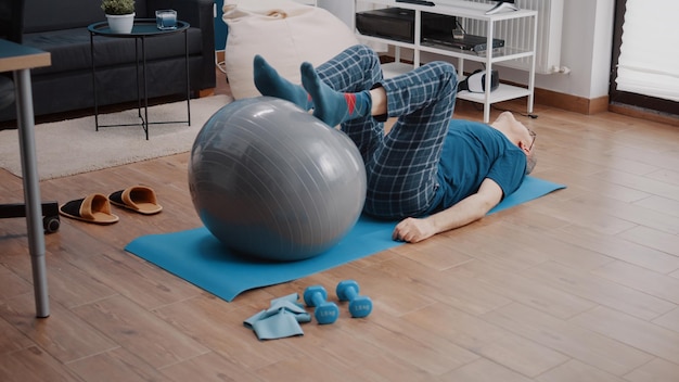 Aged person using toning ball to stretch legs muscles on yoga mat. Senior adult training with fitness equipment, doing physical exercise and activity. Man doing aerobics workout.