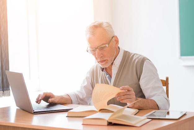 Aged professor male working with laptop while reading book in classroom