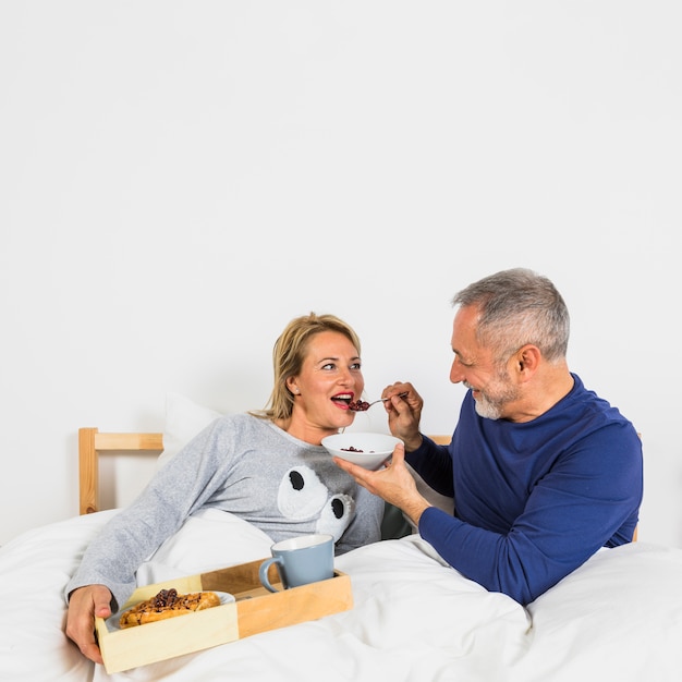 Aged smiling man giving berries to woman in duvet near breakfast on tray on bed