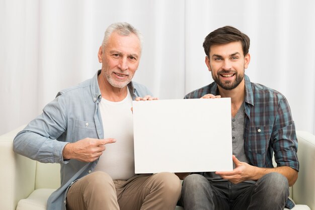 Aged smiling man pointing at paper and young happy guy on sofa