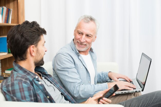 Aged smiling man with laptop and young guy using smartphone on sofa