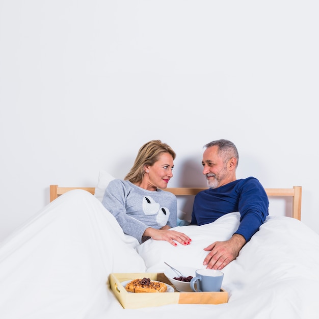 Aged smiling man and woman in duvet on bed near breakfast on tray