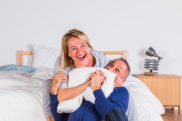 Aged smiling woman hugging man and having fun with pillow near bed