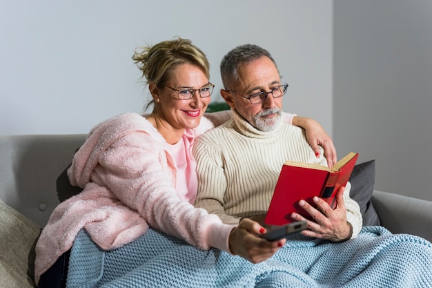 Aged smiling woman with TV remote watching TV and man reading book on settee