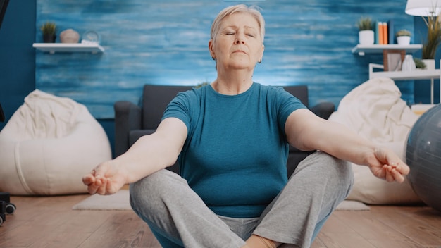 フロアマットの上に蓮華座に座って瞑想をしている老婆。身体運動や体操のトレーニングを行う前に、レクリエーションで瞑想している穏やかな人。ヨガのポーズをしている高齢者 Premium写真