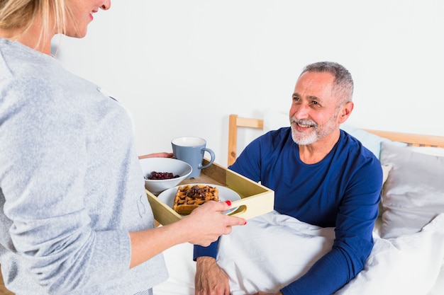 Aged woman giving breakfast to smiling man in duvet on bed