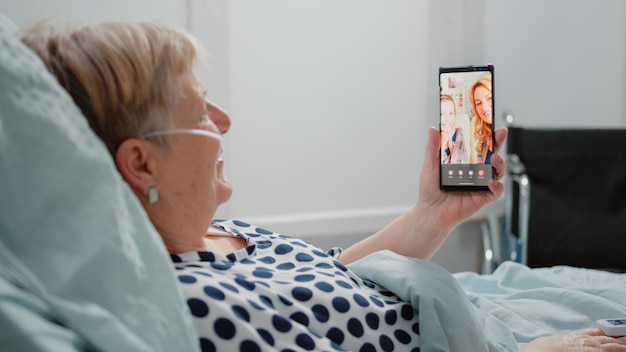 Возрасте женщина, держащая смартфон с видеозвонком для удаленного общения с племянницей и дочерью. Больной пациент использует онлайн-конференцию, чтобы поговорить с родственниками, лежа в больничной койке.