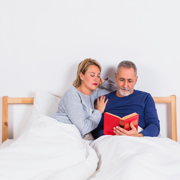 Aged woman near man with book in duvet on bed