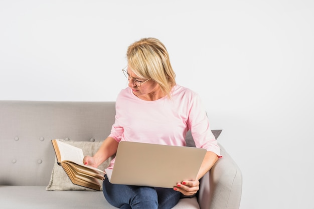 Пожилая женщина в розовой блузке с ноутбуком и книгой на диване
