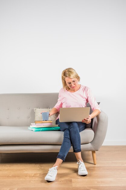 Aged woman in rose blouse with laptop and cup on heap of books on sofa