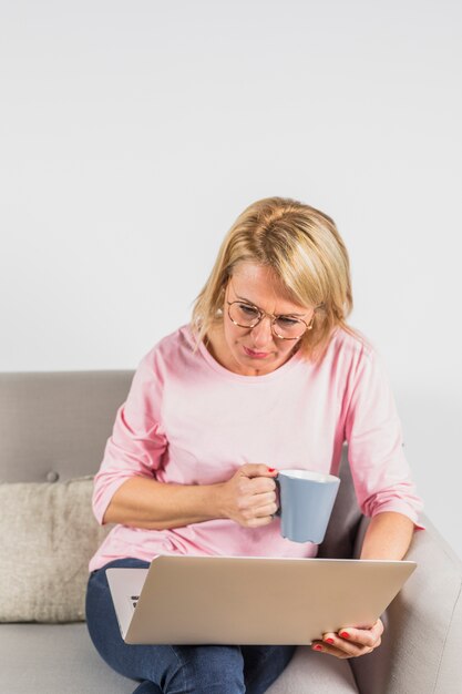 Aged woman in rose blouse with laptop and cup on sofa