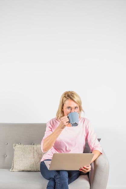 Aged woman in rose blouse with laptop drinking from cup on sofa