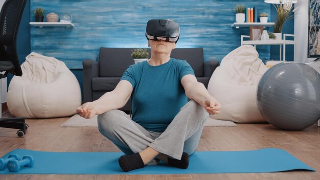 Aged woman using vr glasses to meditate on yoga mat. Elder person wearing virtual reality goggles while doing meditation in lotus position to relax before training. Calm adult with headset