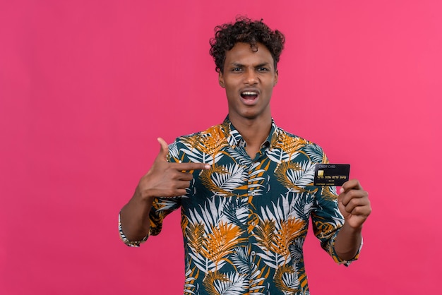 Aggressive and angry young dark-skinned man with curly hair in leaves printed shirt pointing at credit card 