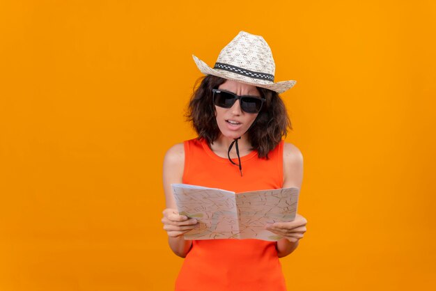 Агрессивная и злая молодая женщина с короткими волосами в оранжевой рубашке в шляпе от солнца и солнечных очках смотрит на карту