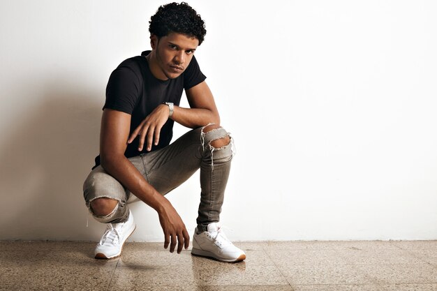 Aggressive and gloomy looking black model in slim torn grey jeans and plain black t-shirt crouching at a white wall