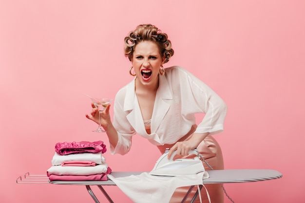 Free photo aggressive housewife with hair curlers holds martini glass and irons white blouse