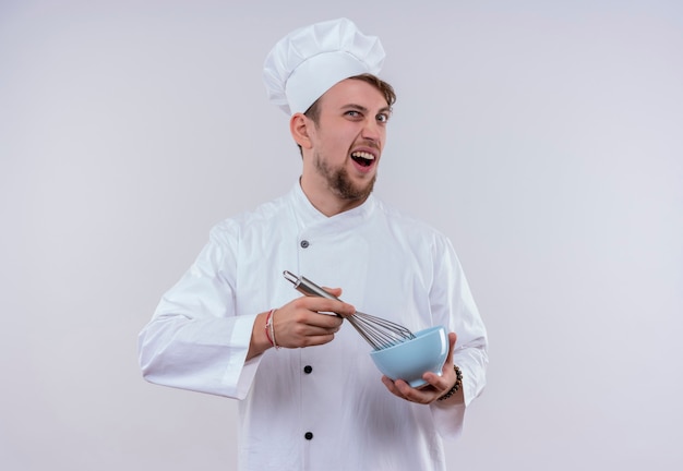 An aggressive young bearded chef man wearing white cooker uniform and hat holding mixer spoon on blue bowl while looking on a white wall