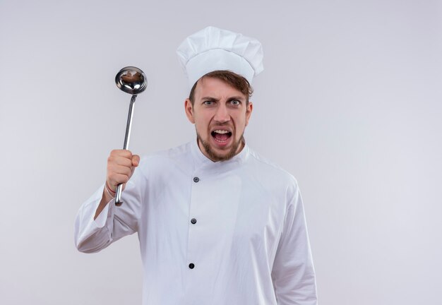 An aggressive young bearded chef man in white uniform holding ladle while looking on a white wall