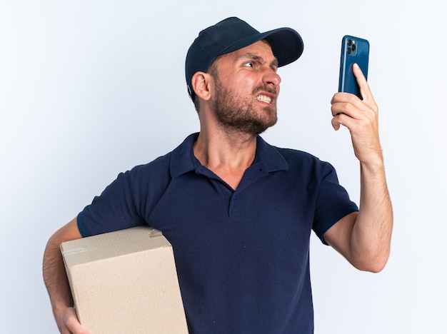 Aggressive young caucasian delivery man in blue uniform and cap holding cardboard box and mobile phone looking at phone isolated on white wall