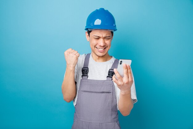 Aggressive young construction worker wearing safety helmet and uniform holding and looking at mobile phone doing yes gesture 