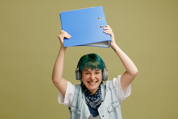 Aggressive young female student wearing headphones and bandana on neck raising folder up looking at camera getting ready to beat someone up with folder isolated on olive green background