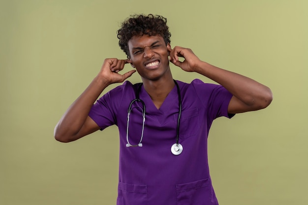 Un giovane medico aggressivo dalla carnagione scura bello con capelli ricci che indossa l'uniforme viola con lo stetoscopio che tiene le mani sulle orecchie mentre su uno spazio verde