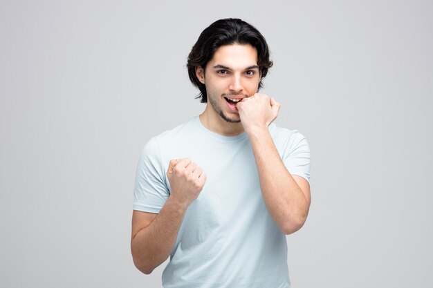 Aggressive young handsome man looking at camera keeping fist in air while biting another fist isolated on white background with copy space