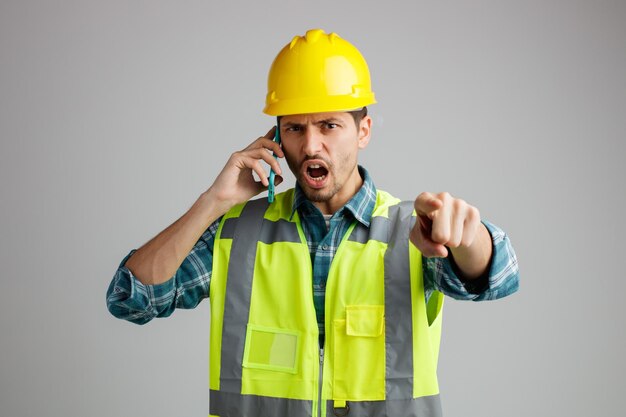 Aggressive young male engineer wearing safety helmet and uniform looking and pointing at camera while talking on phone isolated on white background