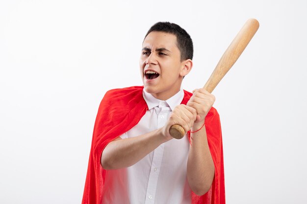 Aggressive young superhero boy in red cape holding baseball bat looking at side getting ready to hit isolated on white background with copy space