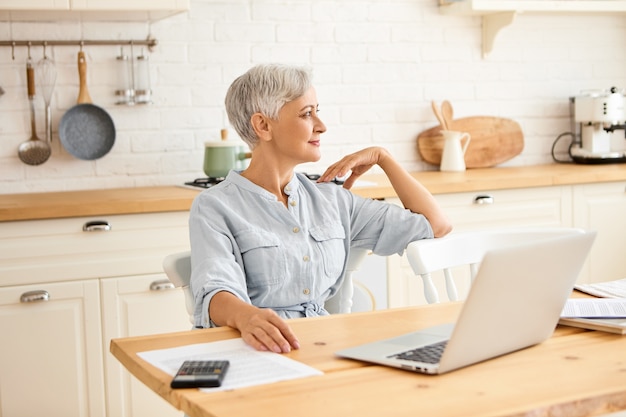 Концепция старения, людей и технологий. Снимок пожилой женщины с короткими волосами в синем платье в помещении, сидящей за кухонным столом с открытым ноутбуком, калькулятором и бумагами и управляющей внутренним бюджетом