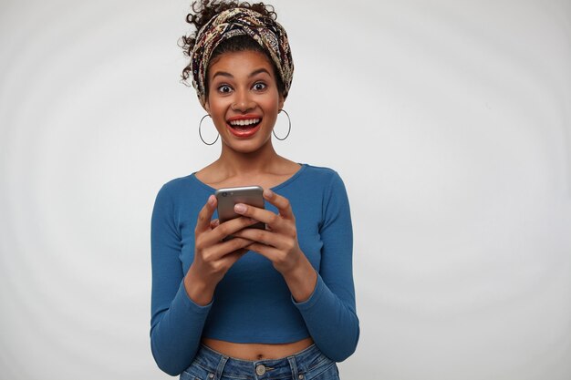 Free photo agitated young attractive dark haired curly female with smartphone in raised hands looking excitedly at camera with broad smile while standing over white background
