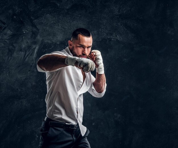 Agressive handsome man in white shirt is demonstraiting his punch at dark photo studio.