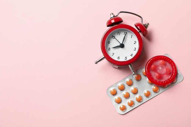 Alarm clock, condom and pills on pink surface