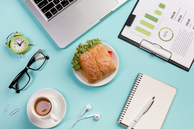 Alarm clock,eyeglasses,breakfast,earphones,spiral notepads and budget plan on clipboard against blue backdrop