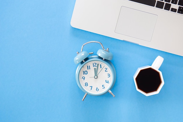 Alarm clock laptop and coffee cup on blue background flat lay