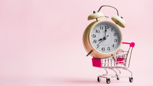Alarm clock in miniature shopping trolley against pink background