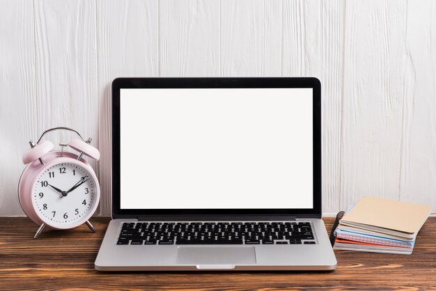 Alarm clock; an open laptop and notebook stacked on wooden desk against wall