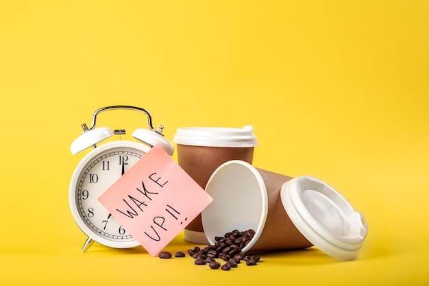 Foto gratuita promemoria della carta della tazza di carta della sveglia e chicchi di caffè su sfondo giallo